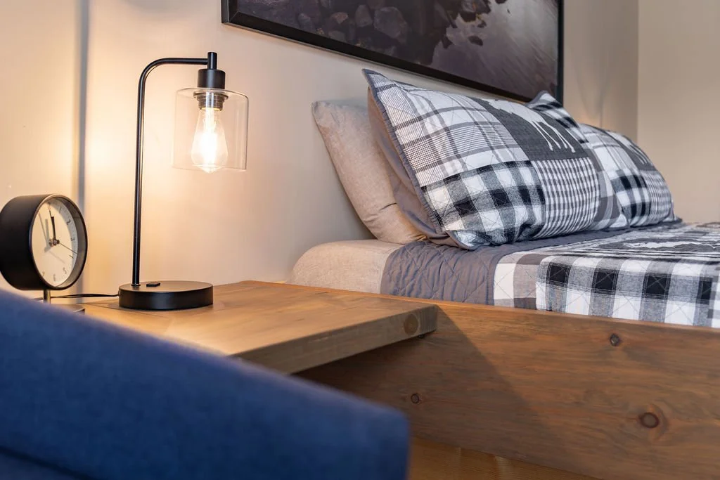 Bedroom with wooden nightstand, table lamp, round clock, bed with plaid-patterned pillows, and abstract wall art.