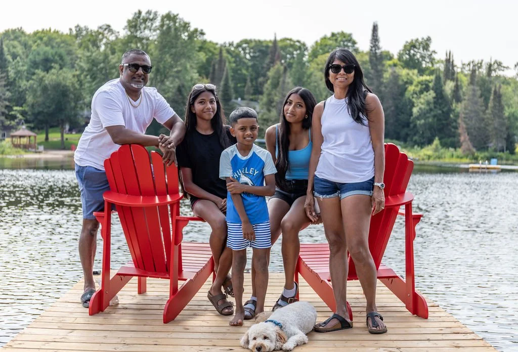 A family of six with three children and two adults, standing on a wooden dock by a lake, with trees and a small pavilion in the background, along with a white dog lying on the dock.