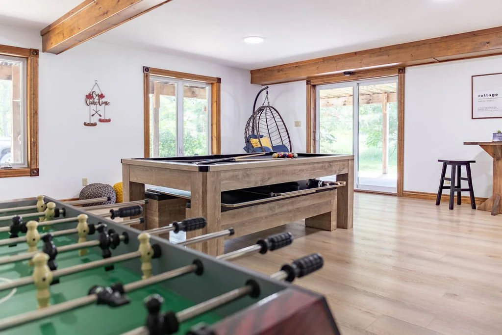 Interior of a recreational room featuring a foosball table in the foreground, a pool table in the center, a hanging egg chair near sliding glass doors, and wooden window frames and beams.
