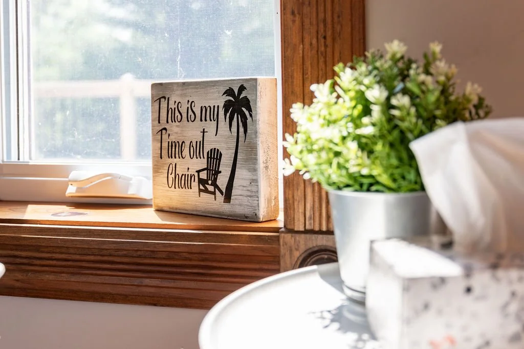 A windowsill with a wooden sign that says "This is my time out chair" next to a tree and palm tree graphic, a white landline phone, and a potted plant with green leaves, with a blurred tissue box in the foreground.