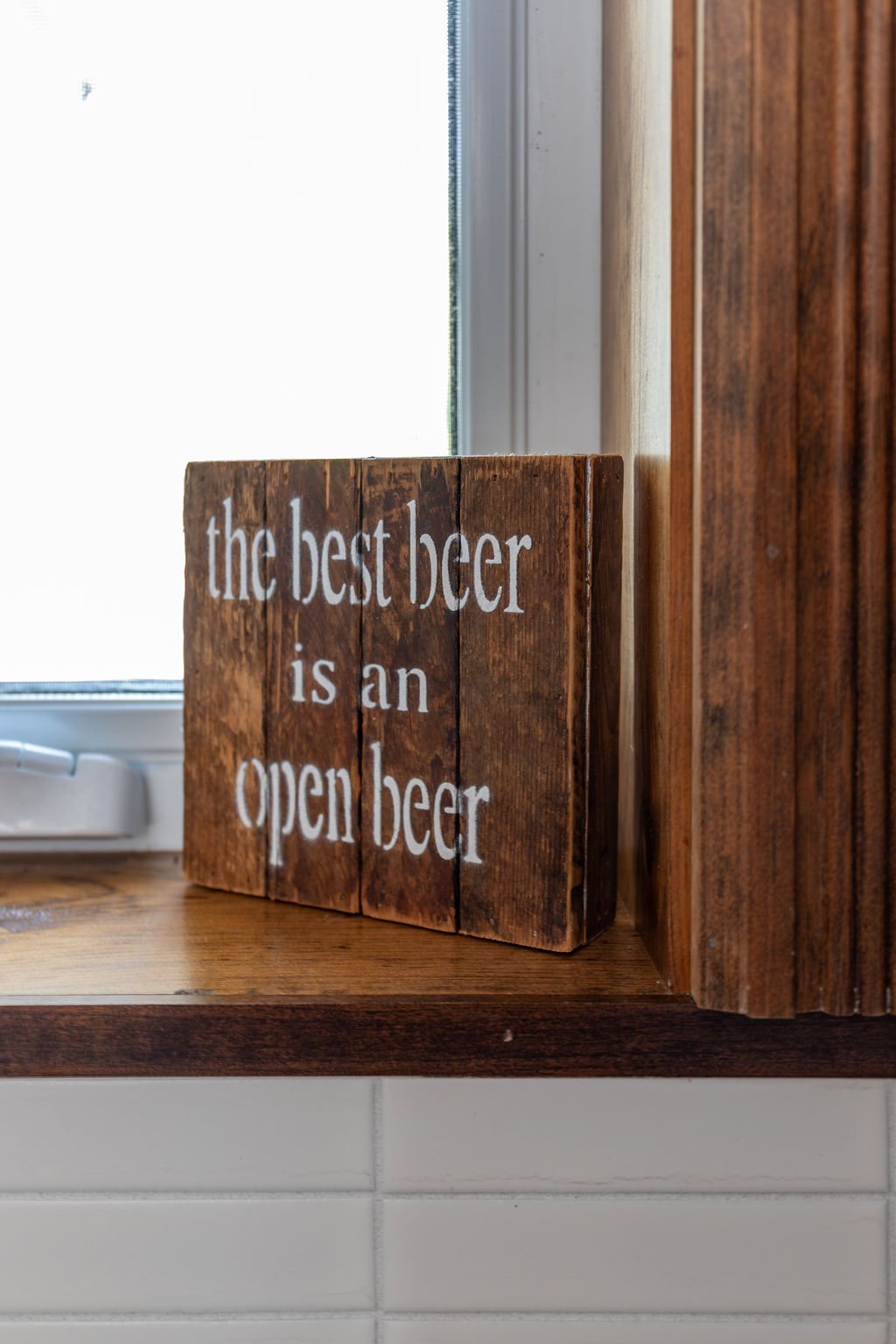 Wooden sign with white lettering that reads "the best beer is an open beer" next to a window and wooden wall.