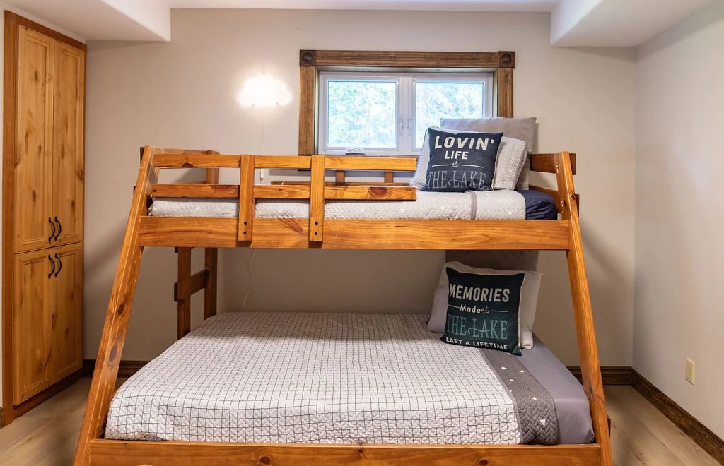 Wooden bunk bed with a mattress and pillows in a small room with a window, a wooden wardrobe, and beige walls.