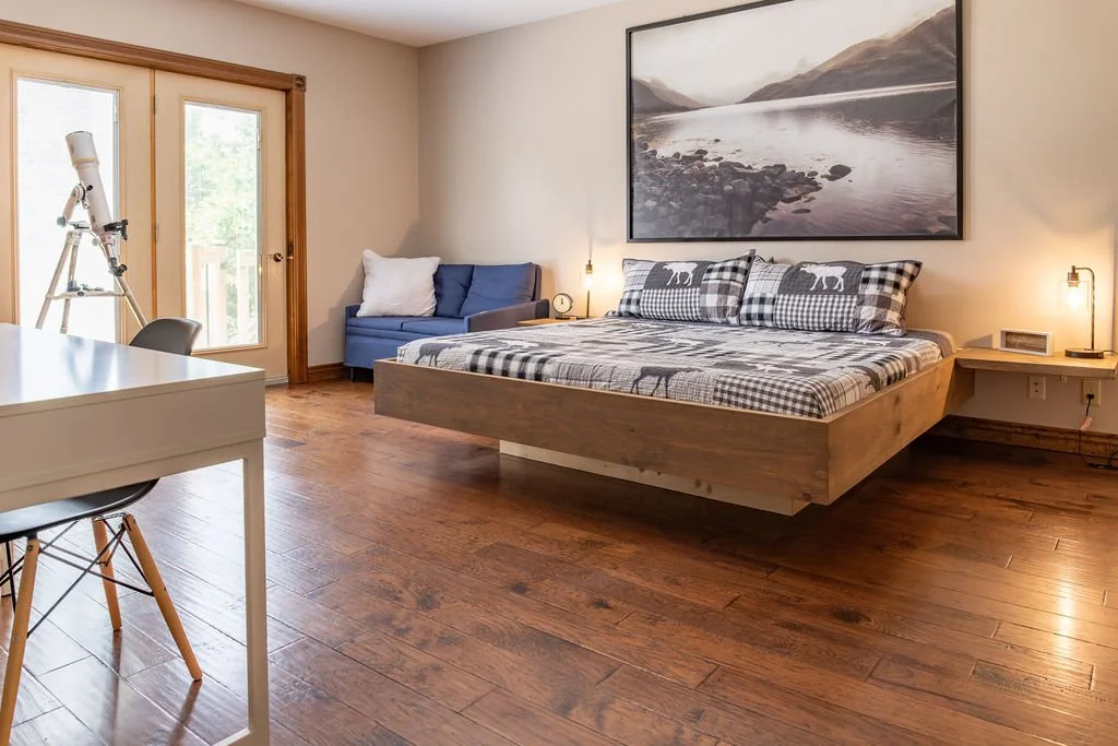 A bedroom with a wooden floor and a large bed with checkered pillows and bedcover featuring bear and plaid patterns. There is a large landscape photograph of a lake and mountains on the wall above the bed. To the right of the bed are wall-mounted lam