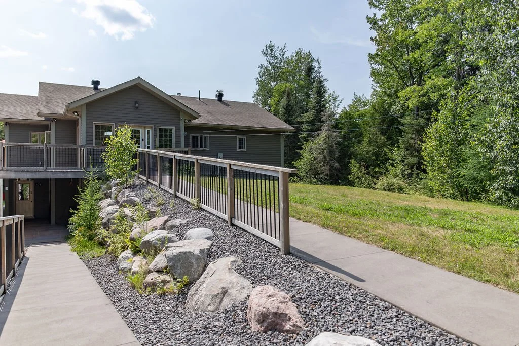 Side view of a house with a deck and a ramp leading down to the yard, surrounded by trees and greenery.