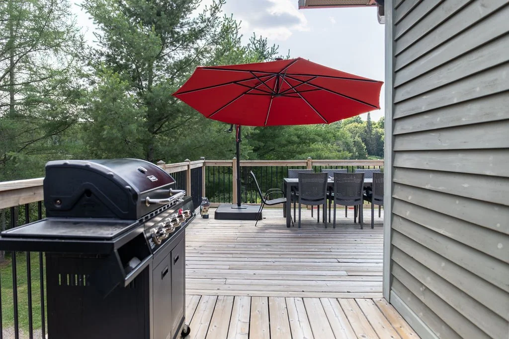 A wooden deck with a black gas grill, a large red patio umbrella, a black outdoor dining table with six matching chairs, and a view of trees in the background.