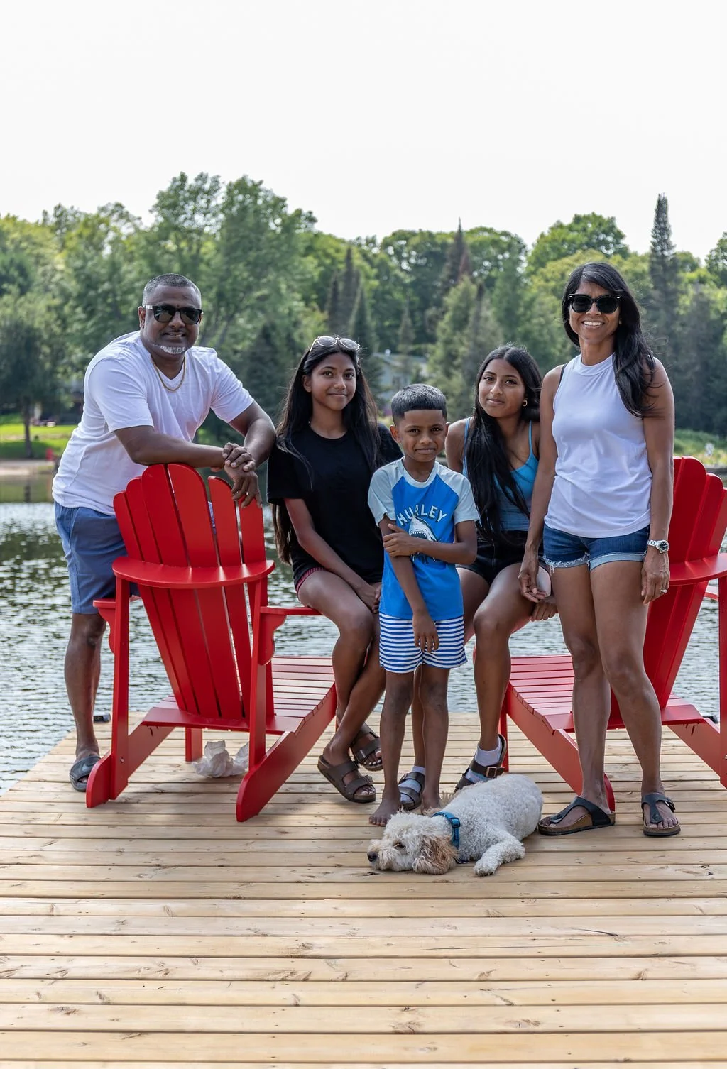 A group of six people, including two adults and four children, standing on a wooden dock by a lake with lush green trees in the background. A white dog is lying on the dock in front of them.