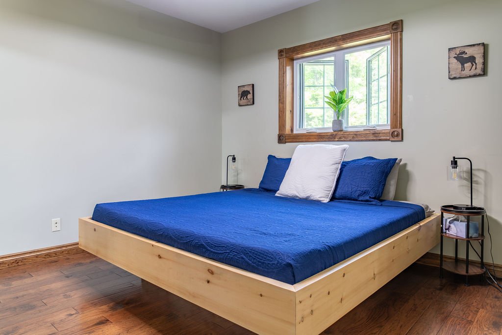 Bedroom with a wooden bed frame, blue bedspread, white pillow, blue pillows, a window with a wooden frame, a potted plant, and two wall art pieces of animals.