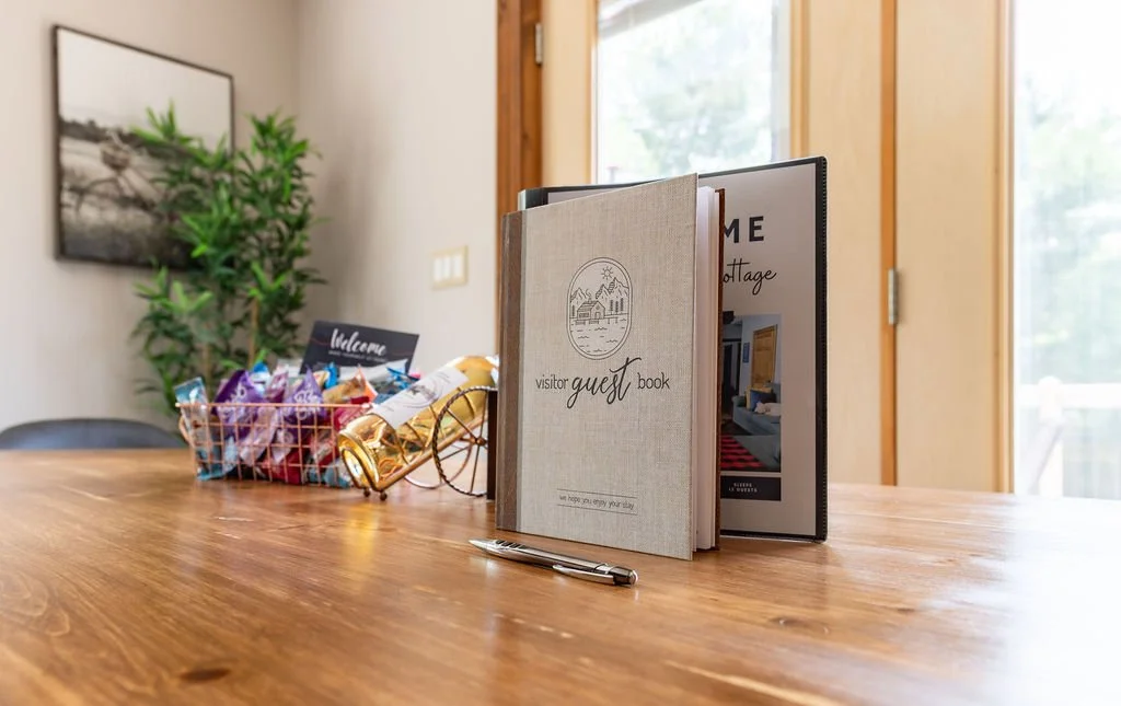 A wooden table with a guest book, a brochure, and a pen in the foreground. In the background, there is a basket filled with snacks, a welcome sign, a potted plant, and a framed picture on the wall. A glass door with a wooden frame is also visible.