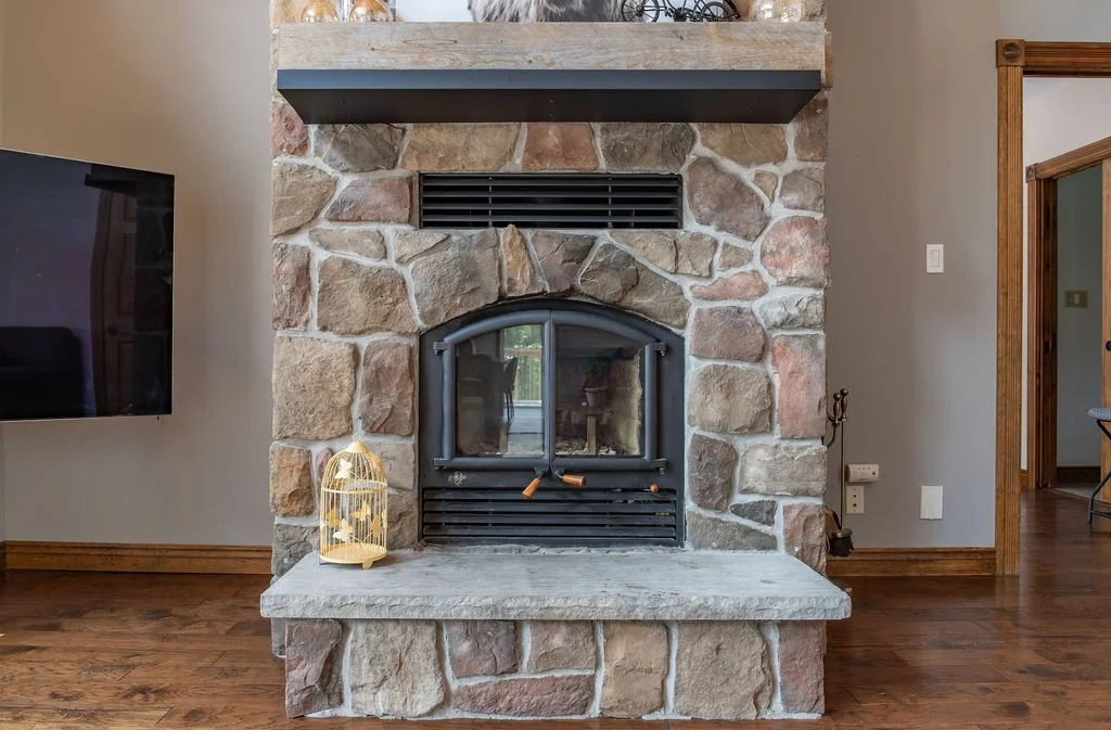 A stone fireplace with a black metal stove inside, a birdcage with a small bird figurine on top, and a wooden mantel with decorative items, in a living room with hardwood flooring.