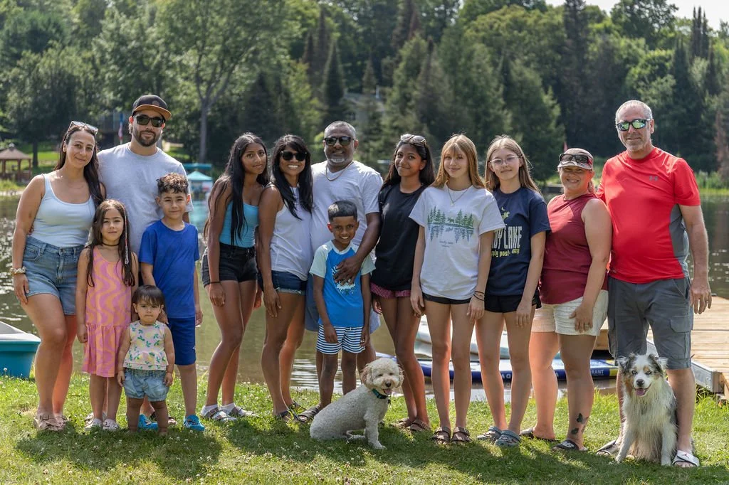 Group of people posing outdoors near a lake, with trees and a dock visible in the background. There are adults, children, and dogs in the photo.