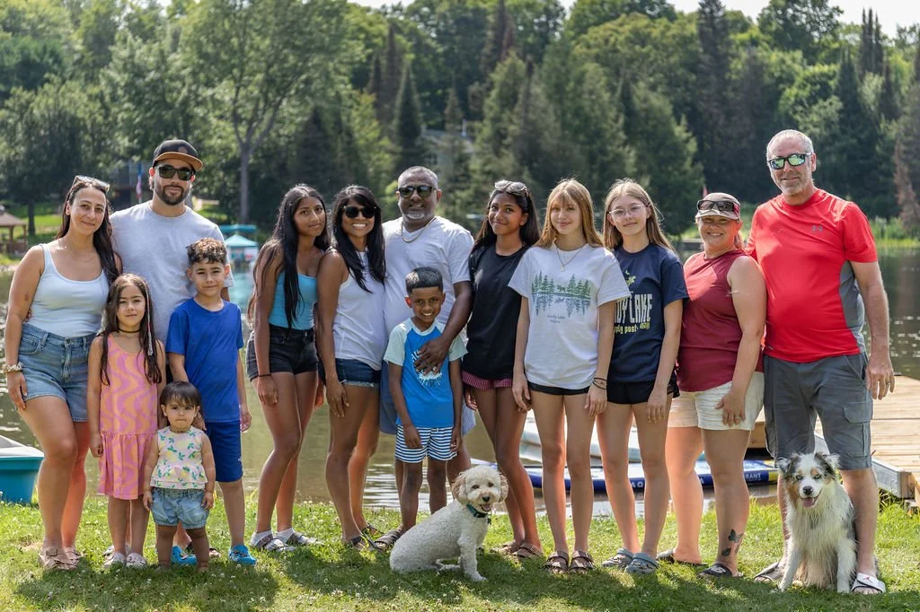 Group of people and dogs outdoors by a lake, with trees in the background.