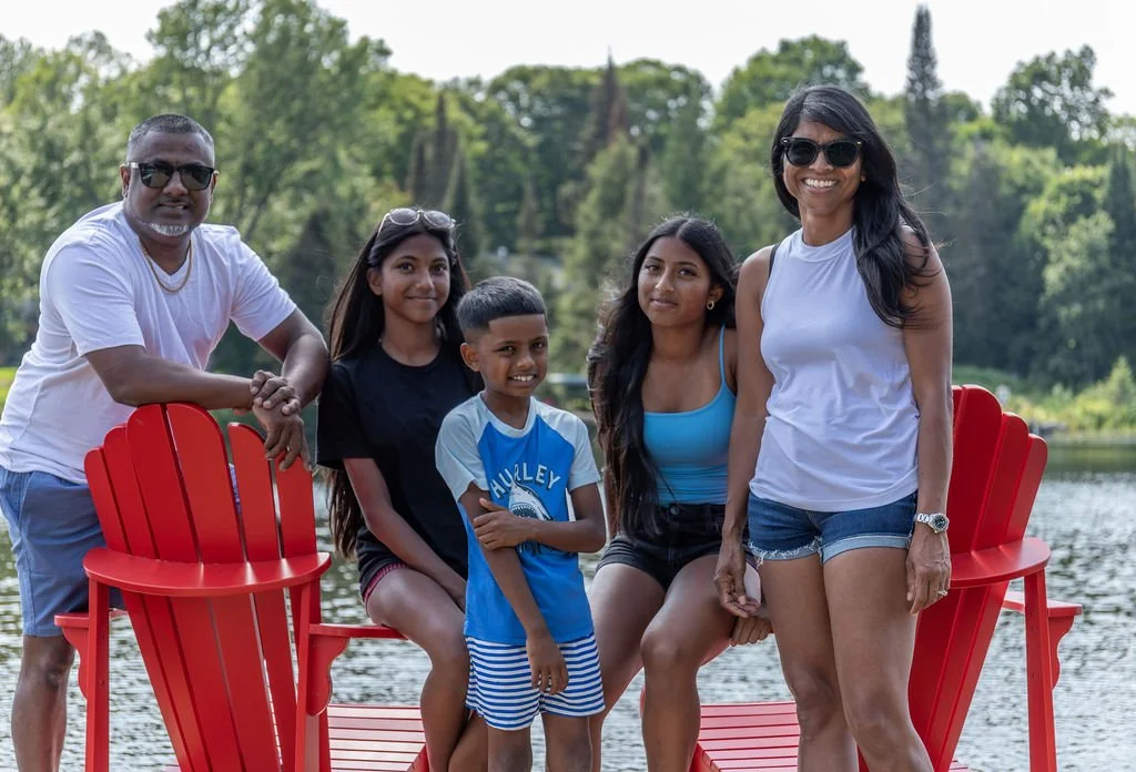 Group of five people, two men and three women, standing by a lake with trees in the background. They are smiling and wearing casual summer clothing. Two red Adirondack chairs are visible, with some people leaning on them.