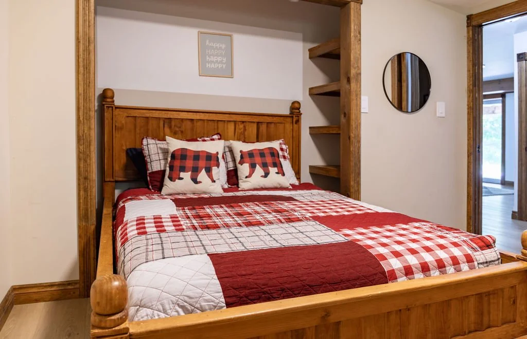A cozy bedroom with a wooden bed frame and plaid bedding, including pillows with bear patterns, in a rustic room with wooden shelves and a round mirror.