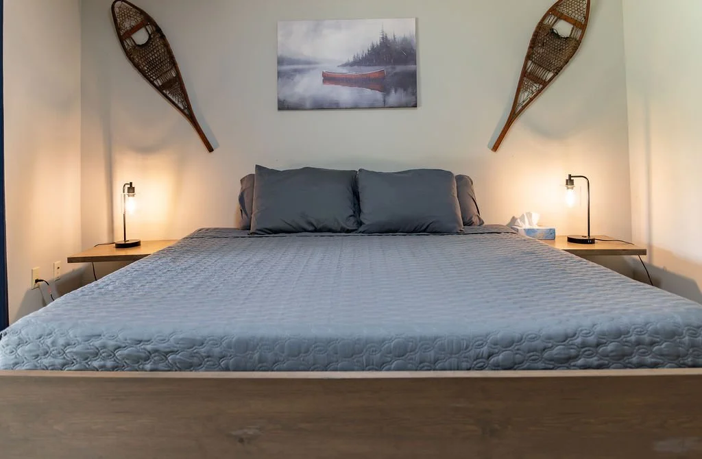 A neatly made bed with gray bedding and matching pillows in a bedroom. On each side of the bed are wooden nightstands with small black lamps. Above the bed is a painting of a canoe on a misty lake, and on the walls, there are two wooden snowshoes han