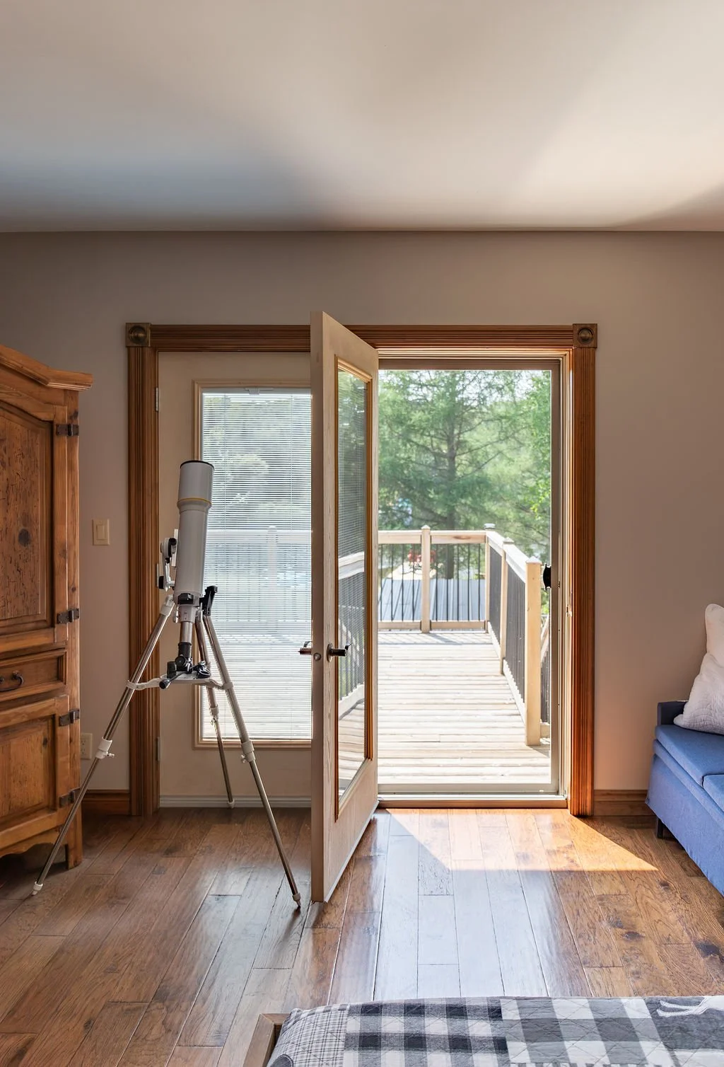 A living room interior with a wooden floor, a wooden armoire on the left, a telescope next to a door, and a couch with a pillow on the right. The door is open, leading to a small balcony with wooden flooring and a black railing, overlooking green tre