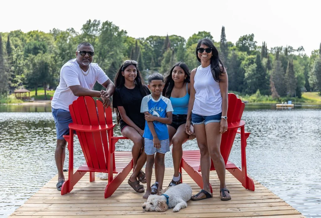 Family of five with two kids, two women, and one man, standing and sitting on a wooden dock by a lake, with a dog lying down in front of them, surrounded by greenery and trees.