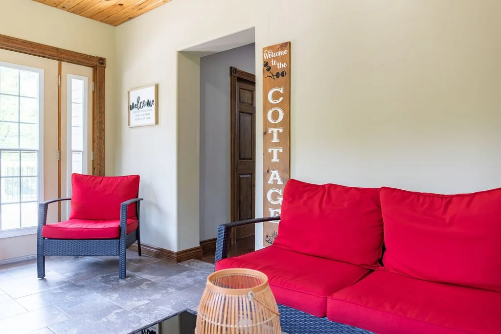 Living room with beige walls, wooden ceiling, red cushioned armchair and sofa, glass door to outside, wooden sign on wall with 'Welcome to the Cottage,' framed artwork, and a wicker lamp
