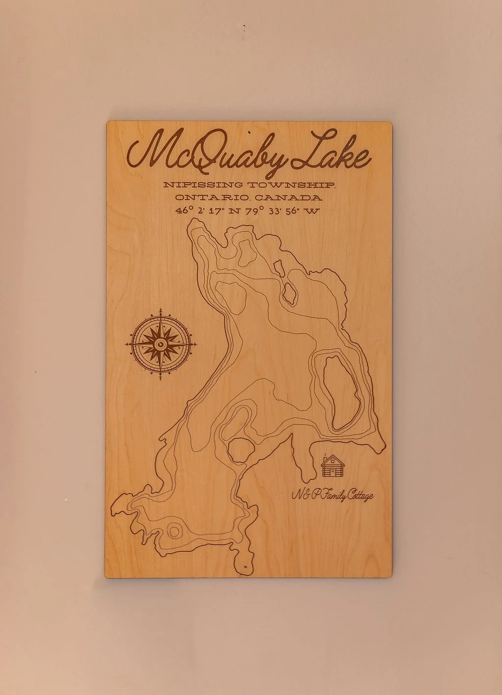Wooden plaque with a topographic map of McQuaby Lake in Nipissing Township, Ontario, Canada. Contains coordinates 46° 2' 17" N, 79° 33' 56" W, a compass rose, and a small drawing of a cottage with the text 'N&W Family Cottage'.