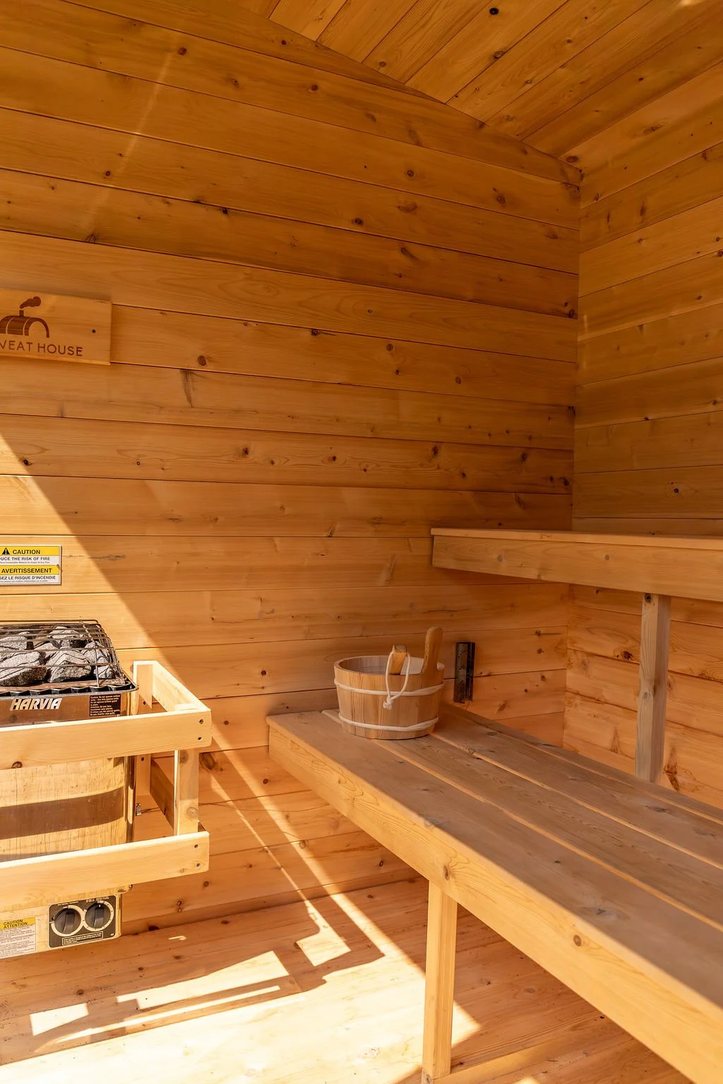 Wood-paneled interior of a sauna room with a wooden bench, a bucket with a ladle, and a small stove for heating rocks, labeled 'Sauna' on the wall.
