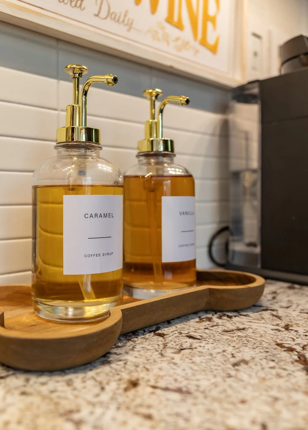 Two glass bottles with gold pumps, labeled 'Caramel' and 'Vanilla', filled with coffee syrup, placed on a wooden tray on a speckled countertop in a cafe.