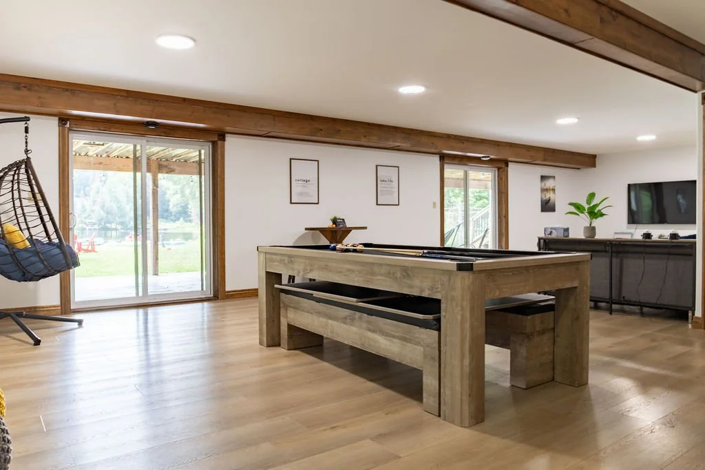 Spacious living room with wooden flooring, large sliding glass doors leading outside, a foosball table in the center, a hanging chair on the left, and a flat-screen TV on the right wall.