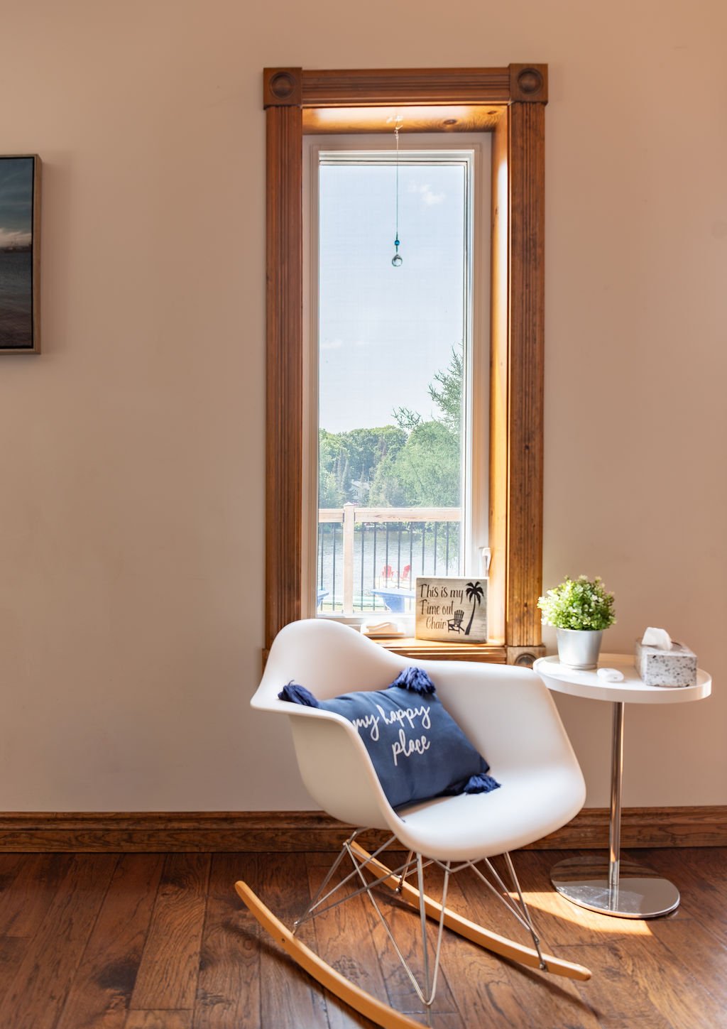 A cozy corner with a white rocking chair that has a blue pillow on it, a small white table with a potted green plant and tissues, a window with a wooden frame, and a lakeside view in the background with trees and water.