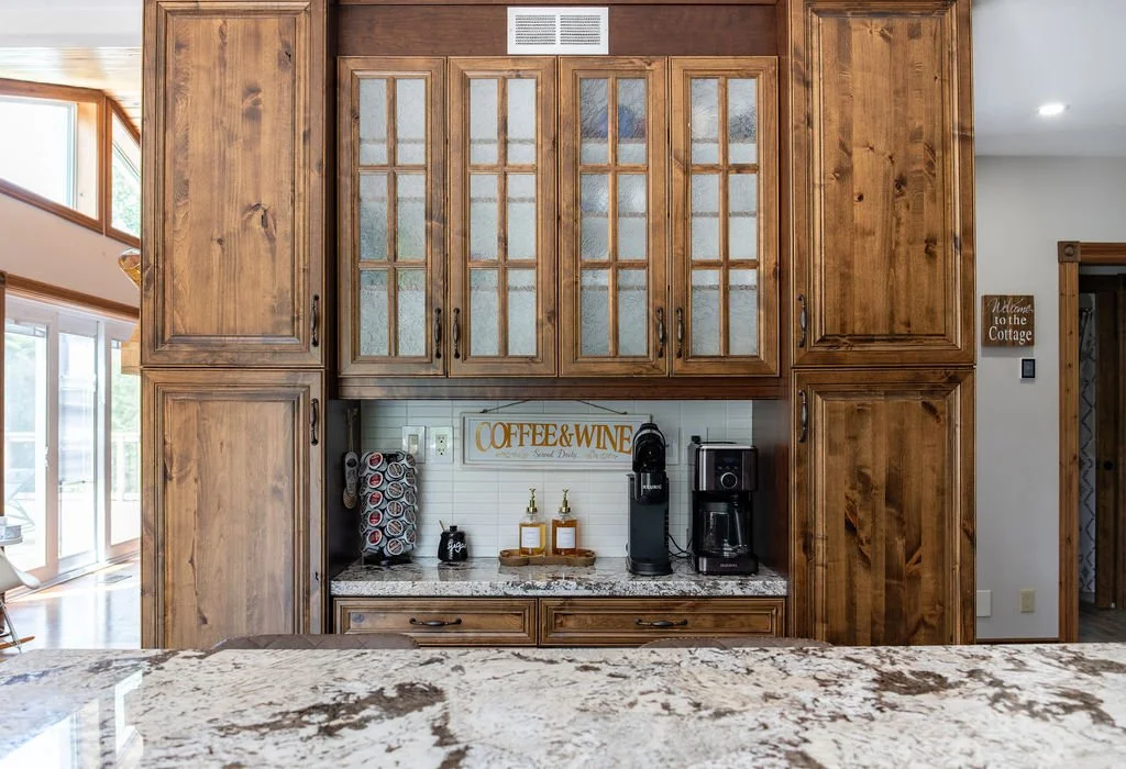 Wooden kitchen cabinetry with glass-paneled doors, a coffee and wine station with a coffee maker, K-Cup holder, and wine bottles, and a granite countertop in a cozy home kitchen.