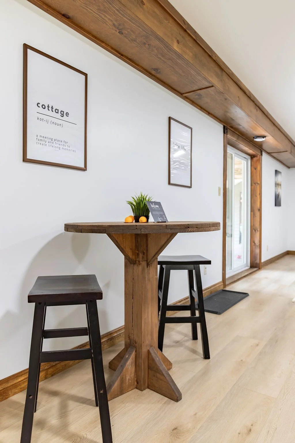 Interior of a cozy room with a wooden table and two black stools, decorated with a small potted plant and a welcome sign. The white wall features framed prints, one defining the word 'cottage.' A glass door leads outside, and a wooden beam runs along