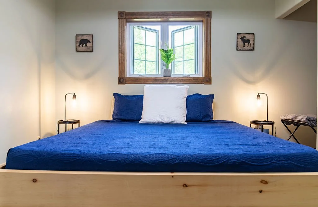 A neatly made bed with a blue quilt and white pillow, two small nightstands with lamps on either side, and window with a wooden frame behind the bed, with deer and bear artwork on the wall.