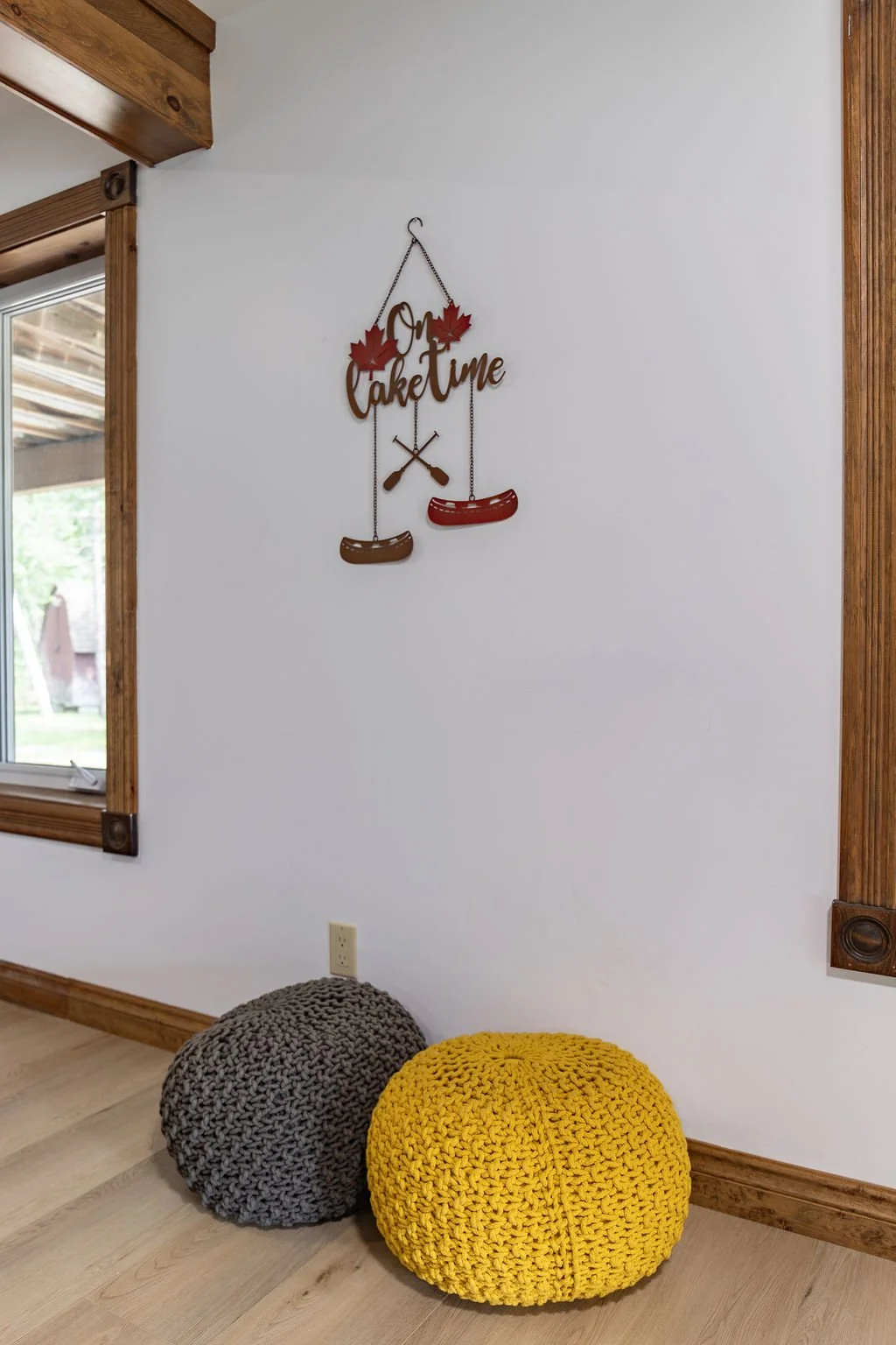 Decorative wall sign reading "Gma Cake Time" with two maple leaves and crossed hot dog icons, next to two knitted poufs in gray and yellow, inside a room with wooden trim and a window.