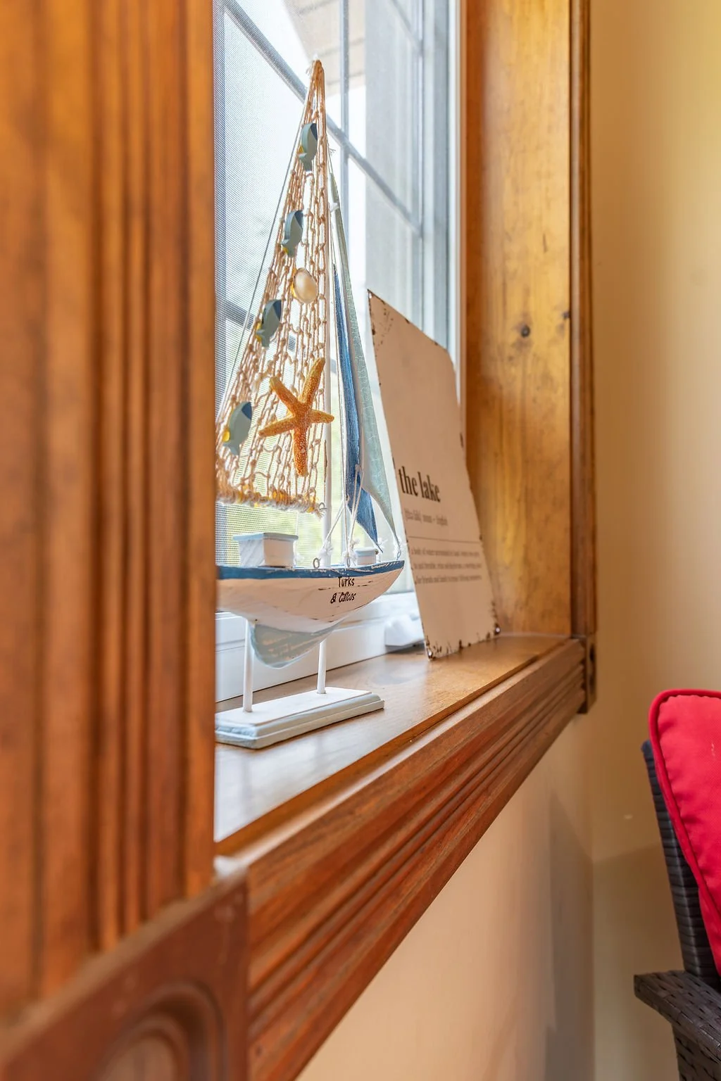 Decorative model sailboat with the words "Turks & Caicos" on the hull, placed on a windowsill next to a sign and a window screen, with wooden framing around the window.