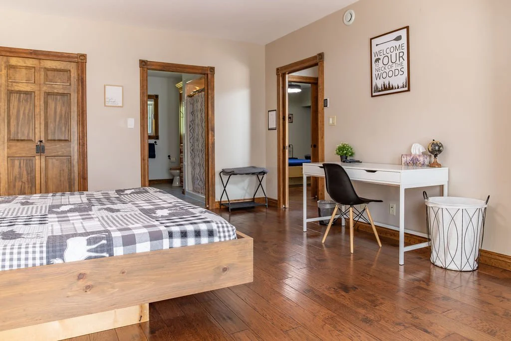Room with a wooden bed, white desk with black chair, wooden closet, and framed wall art that reads 'Welcome to our neck of the woods,' in a cozy, wood-accented space.