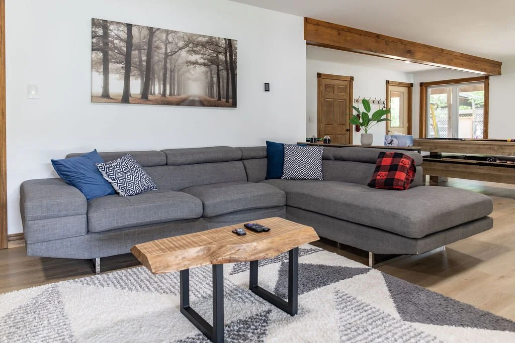 Living room with a gray sectional sofa, patterned and solid throw pillows, a wooden coffee table, a large wall art of a forest, potted plant, and a window with a view of a backyard.