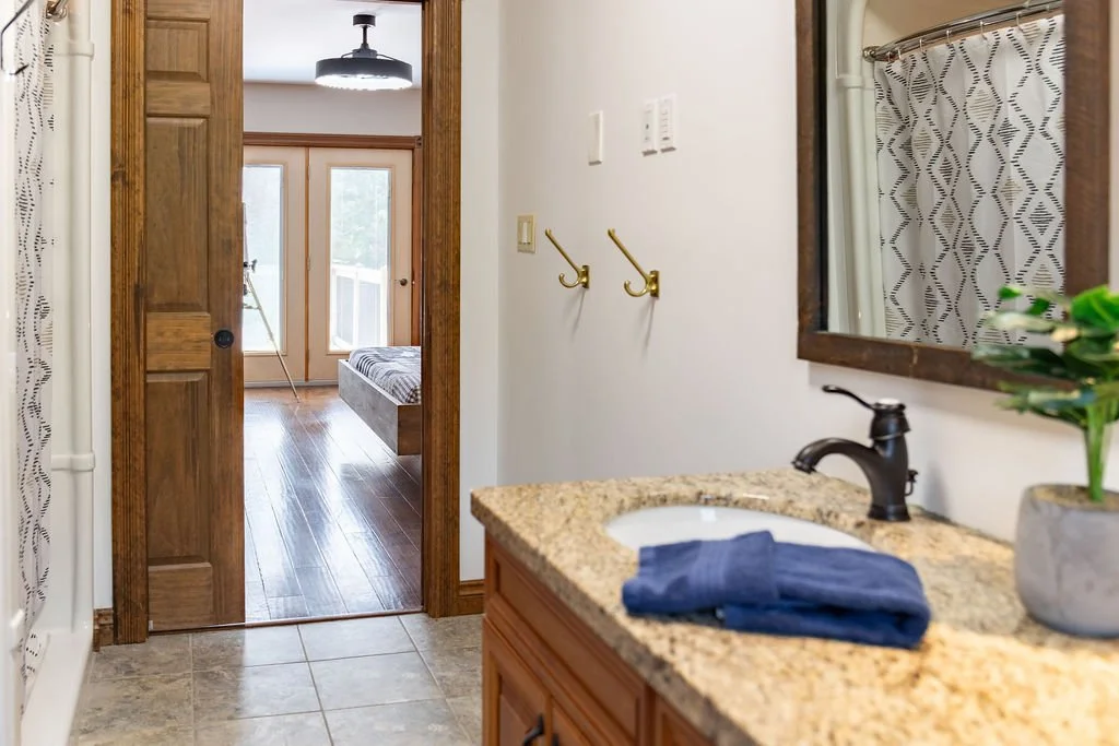 A bathroom with a granite countertop and a sink, a mirror, and a towel. Toward the back, a doorway leads to a room with wooden floors, a bed, and a glass door leading outside. A tripod with a camera or light is visible in the other room.