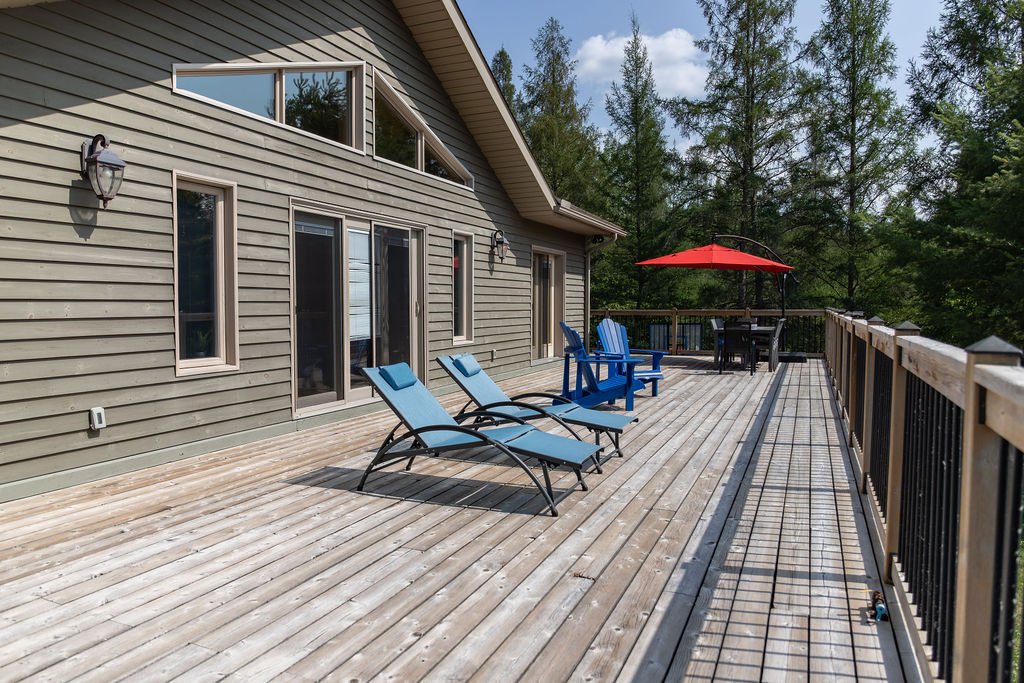 A large wooden deck attached to a house with sliding glass doors and windows, outdoor lounge chairs, a table with a red umbrella, and surrounded by trees.