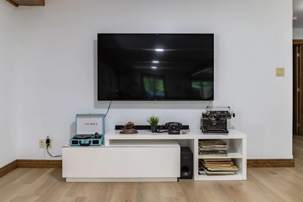 Living room entertainment setup with a large flat-screen TV mounted on a white wall, a white TV stand with a vintage record player, a small plant, a retro telephone, a typewriter, and a collection of magazines.