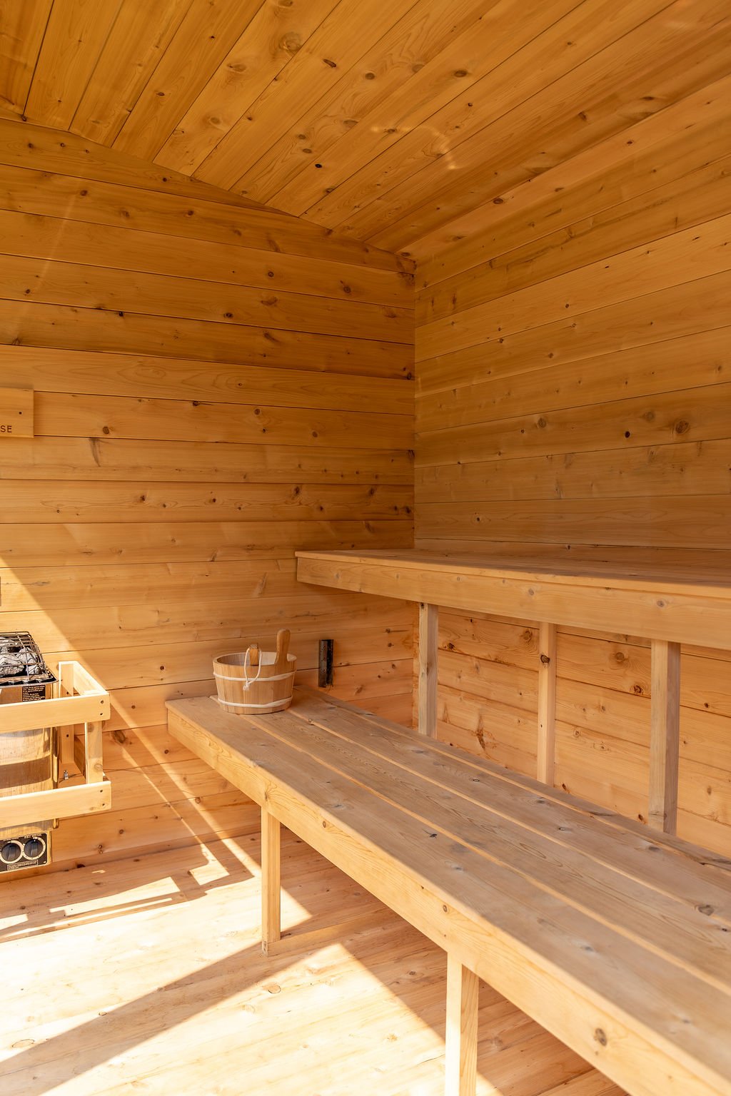 A wooden sauna room with benches, a bucket, and a sauna stove.