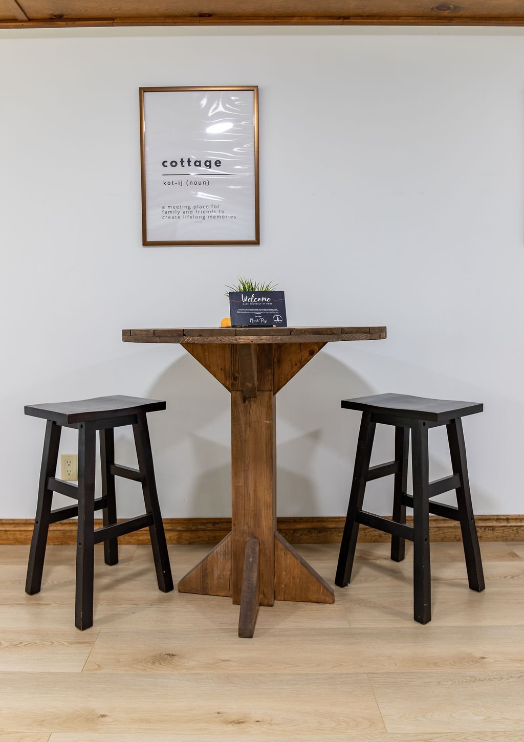 A small wooden table with two black stools against a white wall. A framed sign on the wall defines 'cottage' as a meeting place for family and friends to create memories. A welcome sign and small plant are on the table.