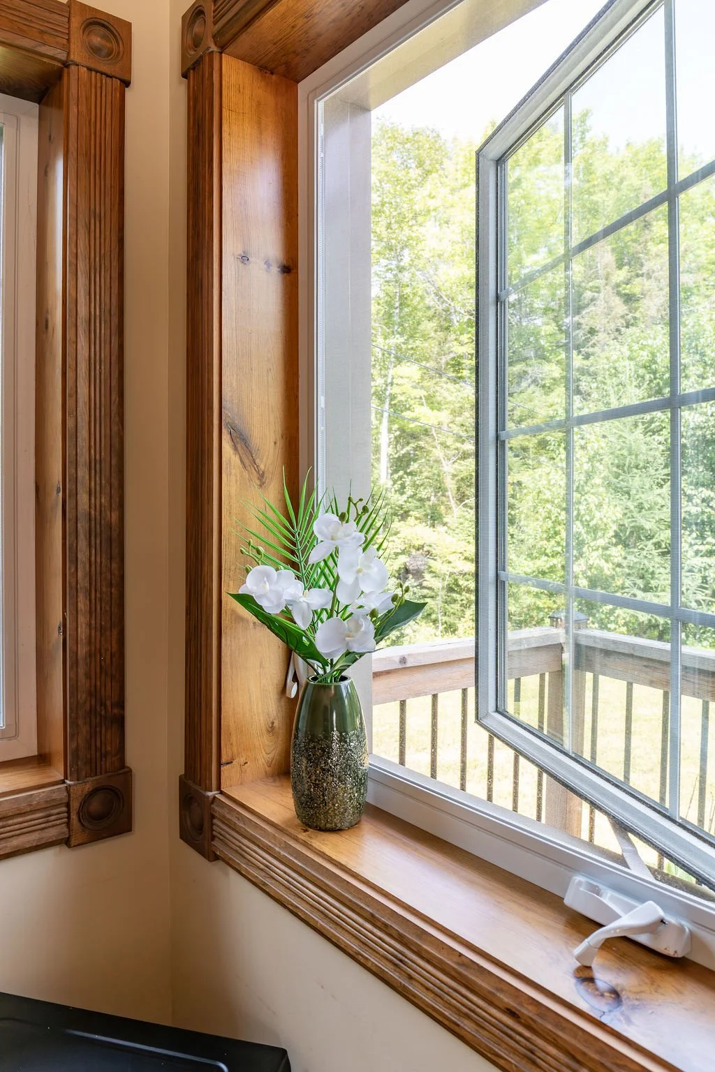 A window with a view of green trees and a wooden deck railing. On the windowsill, there is a green speckled ceramic vase with white flowers and green foliage.