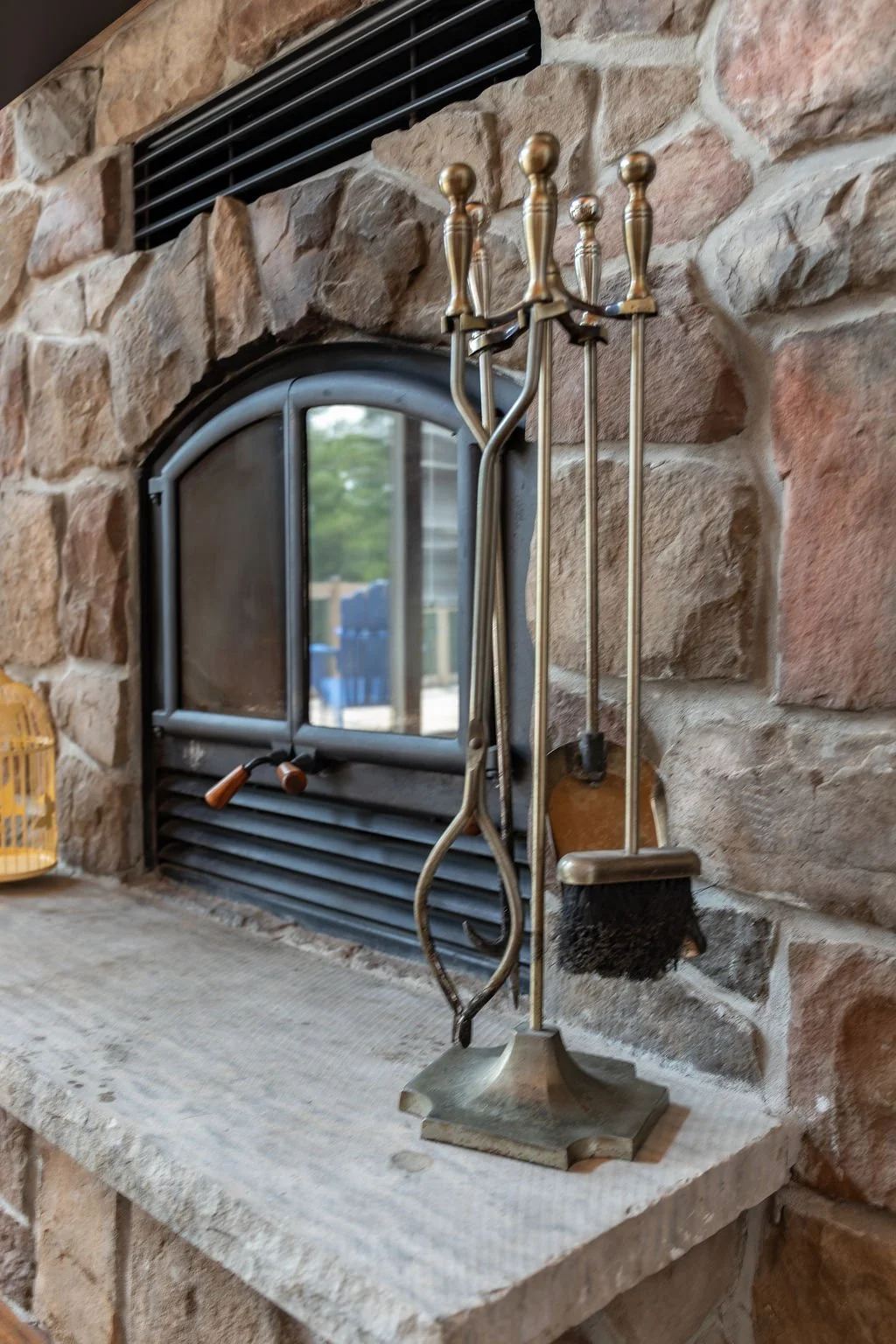 A set of fireplace tools including a poker, tongs, brush, and shovel hanging on a brass stand in front of a stone fireplace.