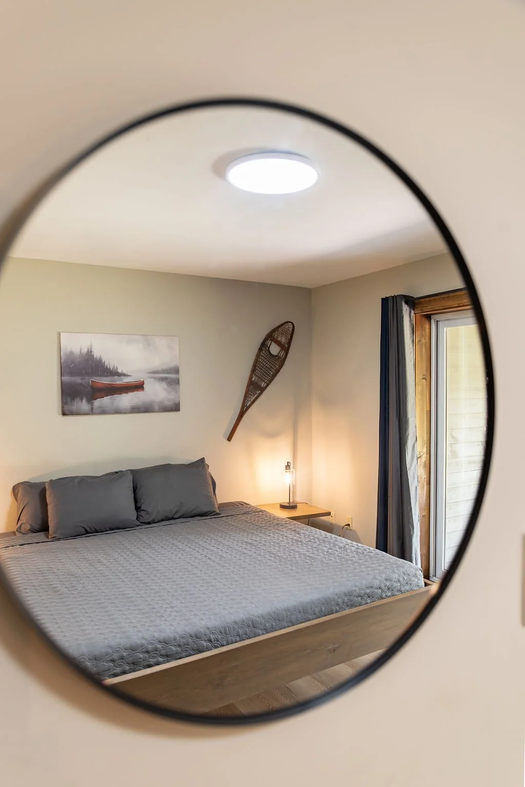 View of a bedroom seen through a circular mirror, showing a bed with gray bedding and pillows, a nightstand with a lamp, a window with curtains, a painting of a boat on water, and snowshoes hanging on the wall.