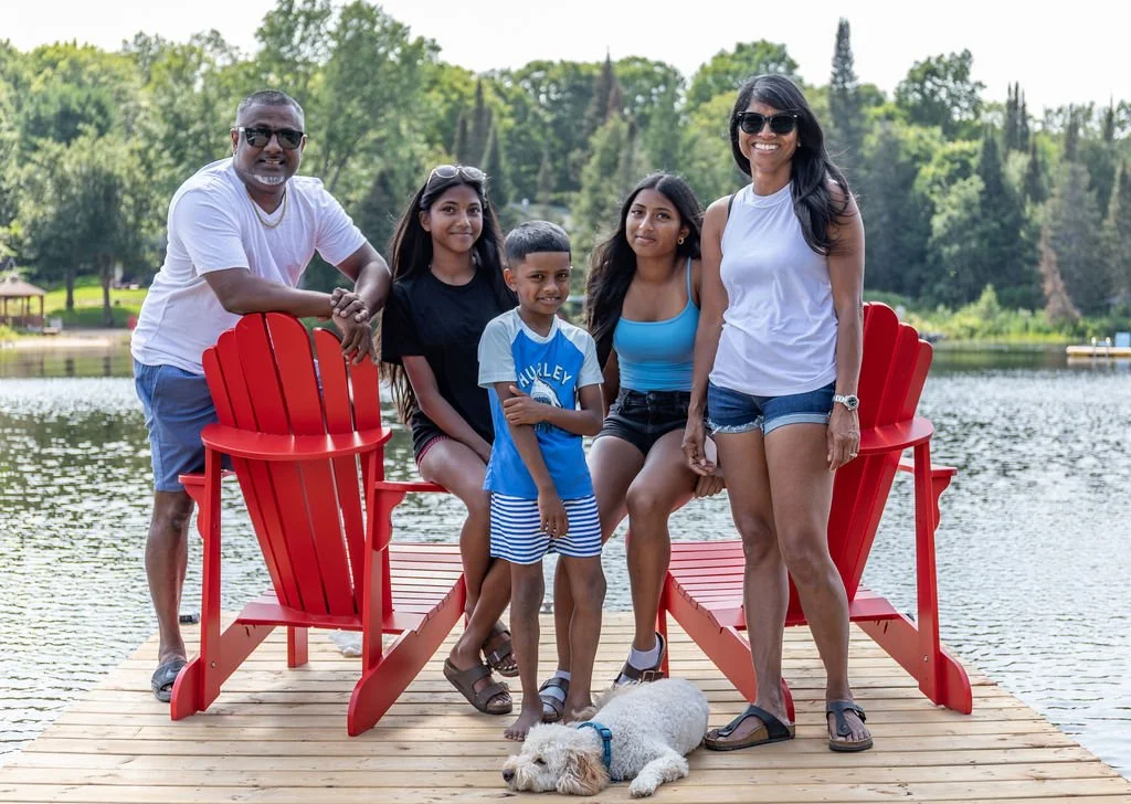 Family of five, including two women, two girls, and a boy, posing on a wooden dock with a dog in front, by a lake with trees in the background.