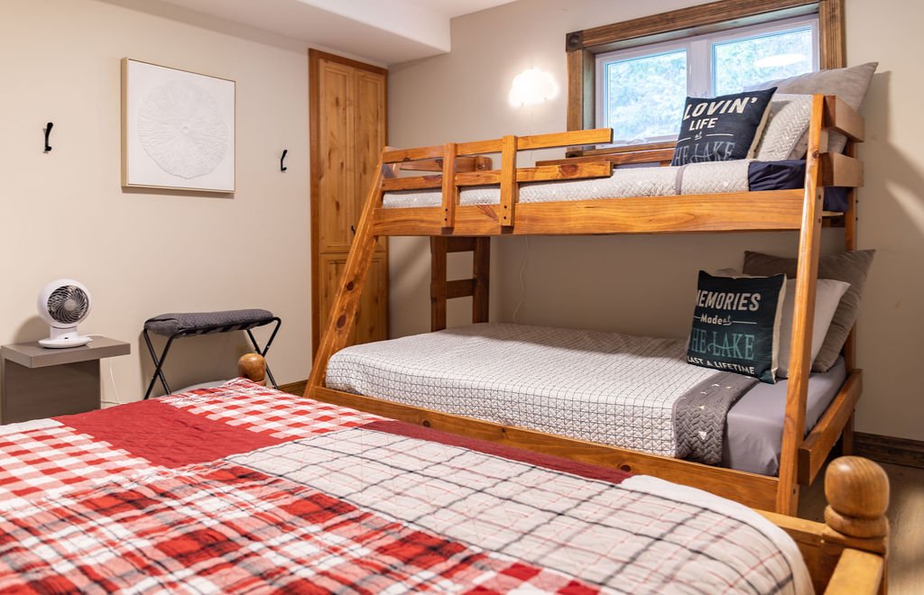 A bedroom with a top bunk bed made of wood, a window above it, and a bed in the foreground with a red plaid blanket. There is a small white fan on a gray nightstand, a folded towel on a black luggage rack, and wall hooks with a white artwork on the w