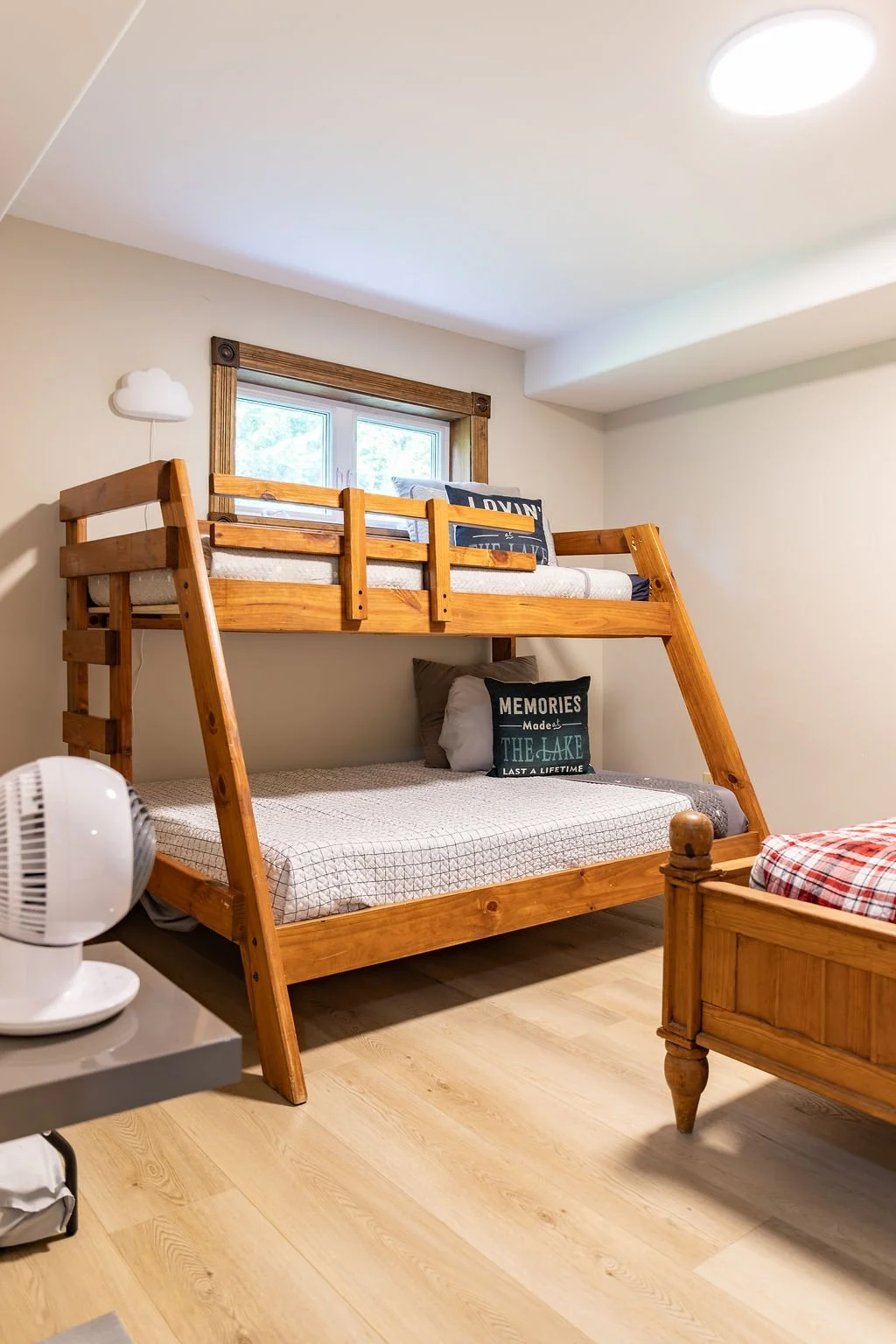 A bedroom with a wooden bunk bed, a window behind the top bunk, and decorative pillows on both bunks. There is a small fan on a gray table to the left and a portion of an adjacent wooden bed with a plaid blanket to the right. The room has light-color