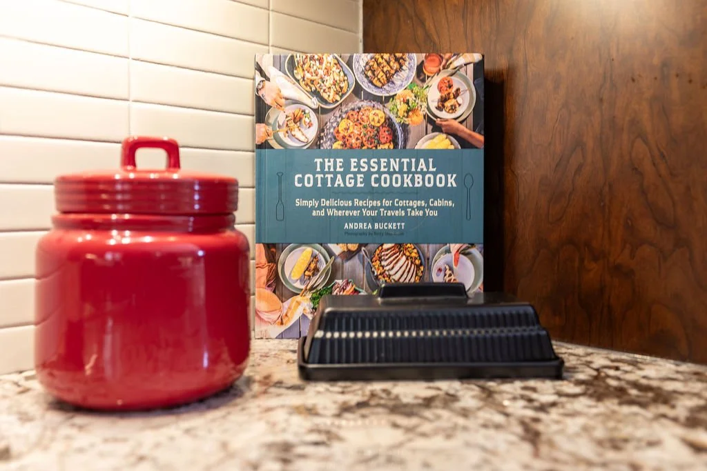 A cookbook titled "The Essential Cottage Cookbook" standing upright on a kitchen counter, with a red ceramic jar with a lid and a black plastic condiment holder in front of it, against a background of white subway tiles and dark brown wood paneling.