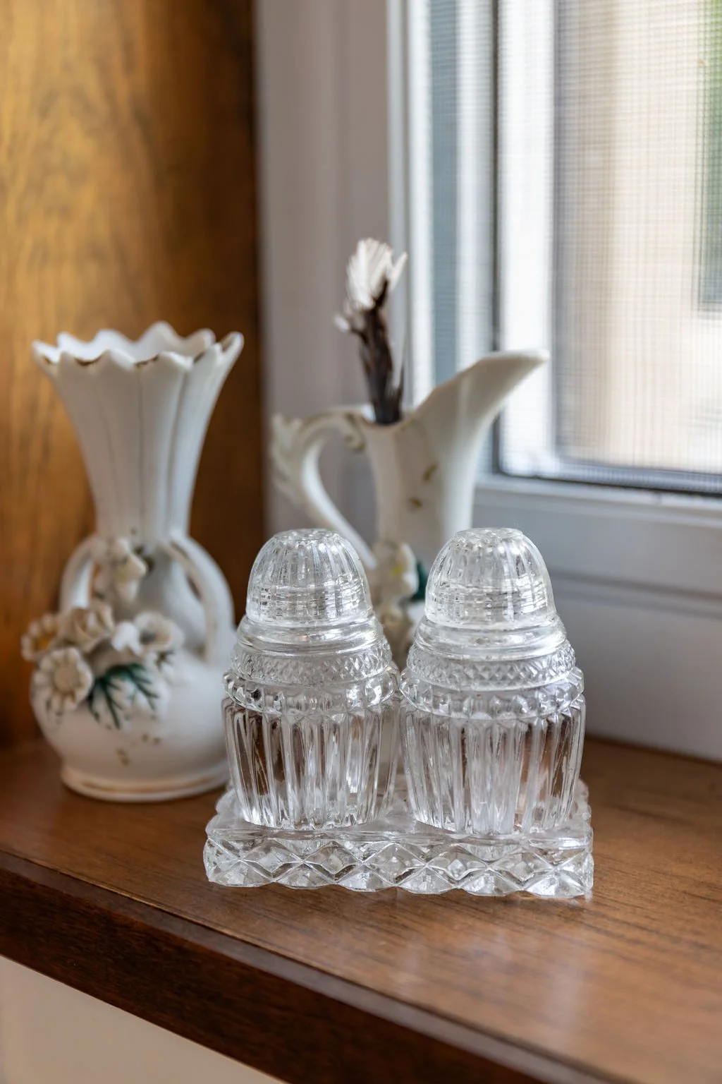 Decorative porcelain vases and a crystal salt and pepper shaker set on a wooden surface near a window.