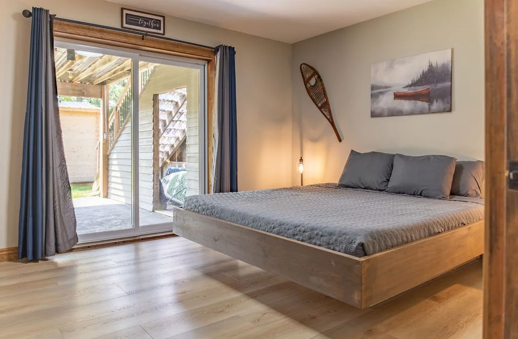 Bedroom with a wooden bed frame, gray bedding, dark gray pillows, sliding glass door with blue curtains, a painting of a boat on the water, and a vintage snowshoe hung on the wall.