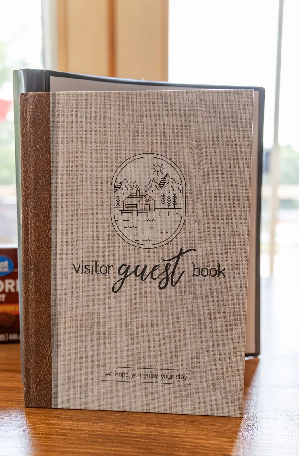 A guest book with a rustic, canvas cover featuring a mountain house scene, trees, water, and a sun, with text that says "visitor guest book" and a message that reads "we hope you enjoy your stay".