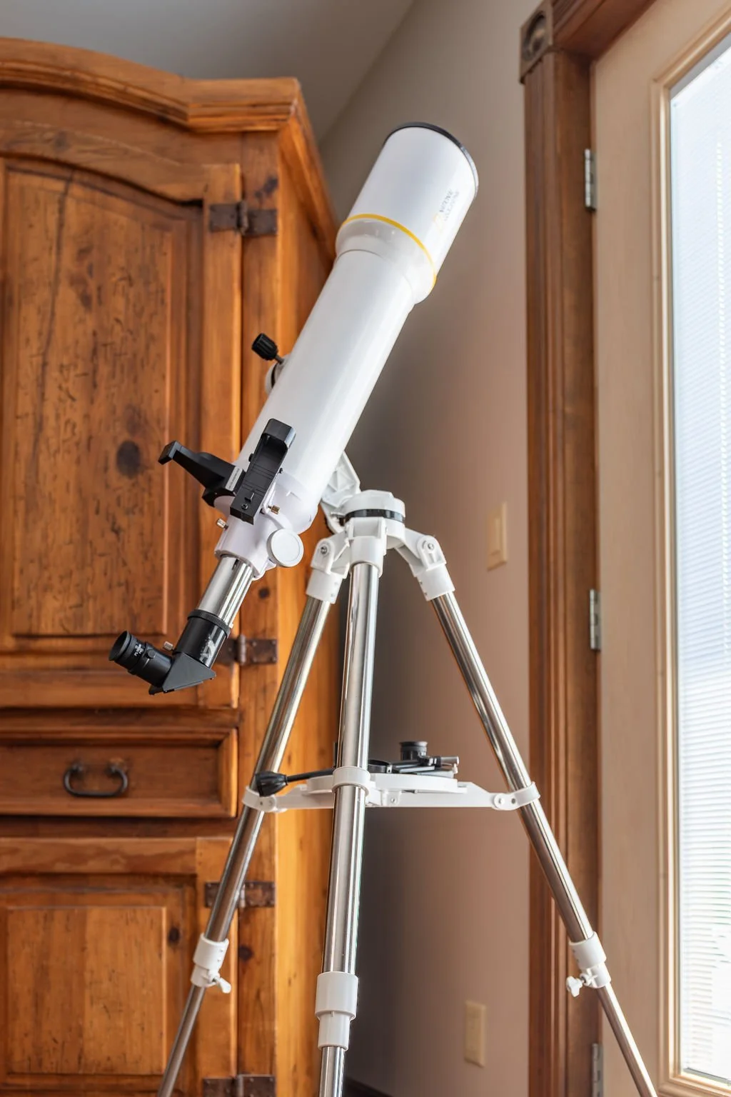 A white telescope on a tripod stands inside a room near a wooden cabinet and window.