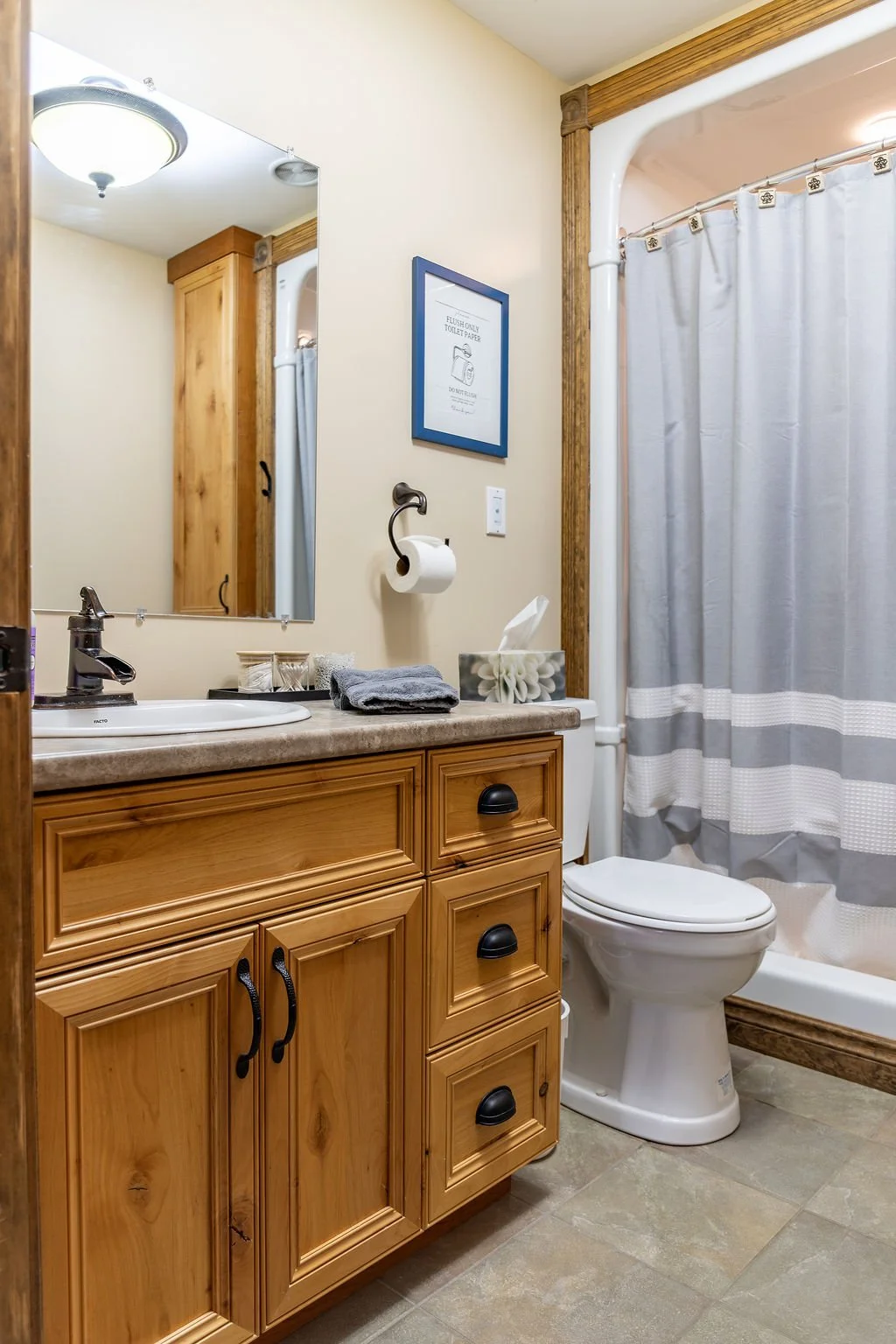 A small bathroom with a wooden vanity, a white sink, a mirror, and a toilet. There are towels, toiletries, a tissue box, and a framed sign on the wall. A shower with a gray and white striped curtain is visible.
